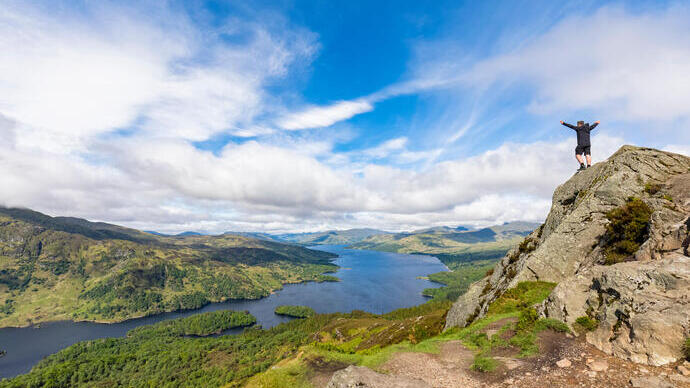 Expansive view of a lake and green hills under a blue sky, with a person standing on a rocky peak, arms raised in triumph.