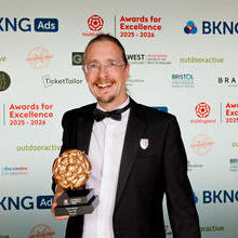 Person in formal attire holding a trophy at an awards event with branded backdrop for Awards for Excellence 2025-2026.