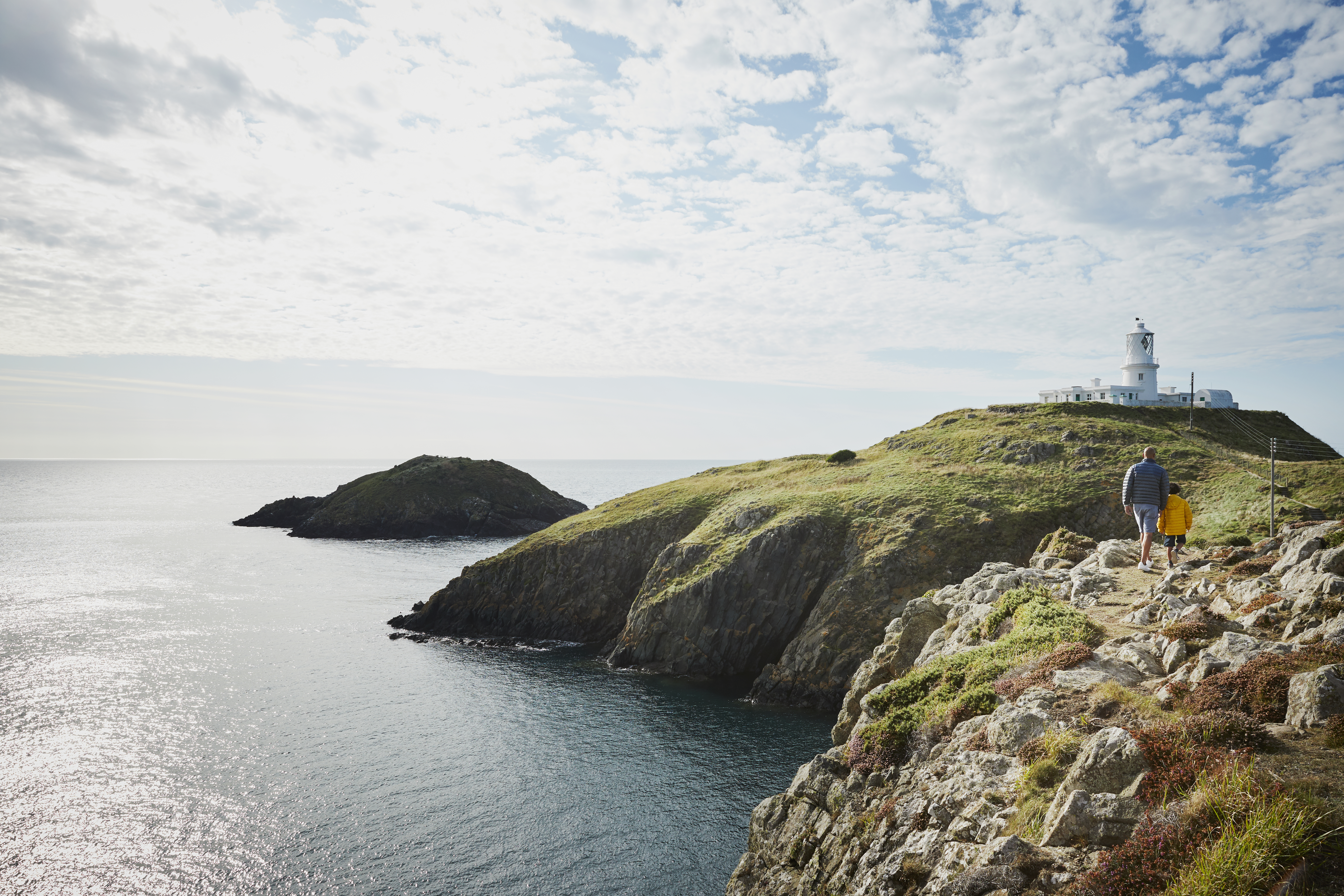Man and child walking on a coastal path towards a lighthouse