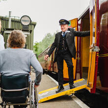 Guard stands on a ramps and leans out of heritage train carriage greeting woman and man using a wheelchair