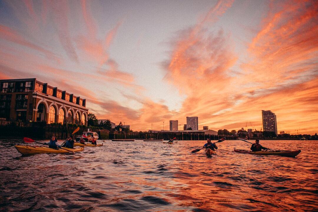 People kayaking on a river against a red sunset sky and city buildings.