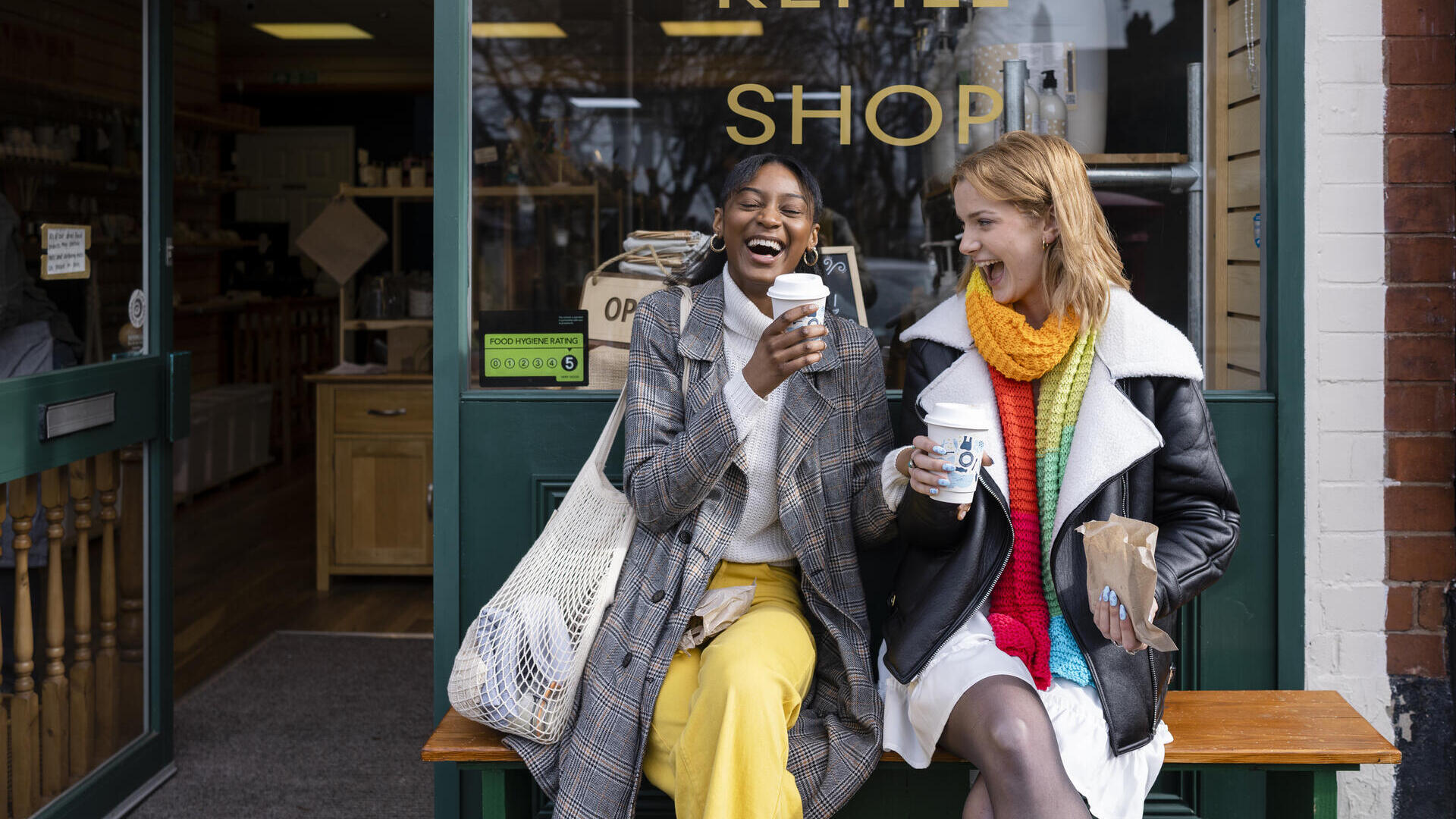 Zwei junge Frauen sitzen auf einer Bank vor einem Café mit Kaffee zum Mitnehmen