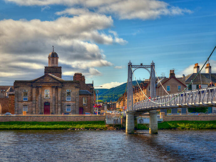 A suspension bridge with people walking across spanning a wide river with houses on the other side