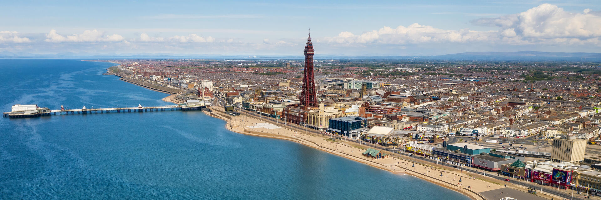 Aerial view of a coastal city with a pier, ferris wheel, tower, sandy beach, and expansive waterfront with cityscape in the background.