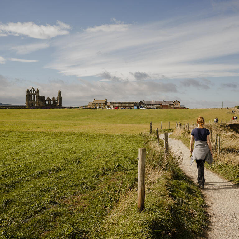 Woman walking along a coastal path towards a ruined abbey