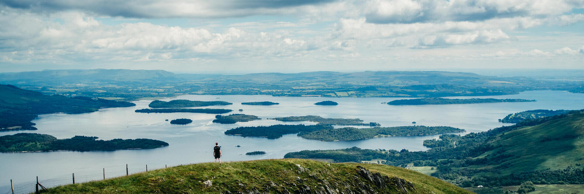 Amplia panorámica de un excursionista de pie en lo alto de una colina contemplando praderas y lagos.