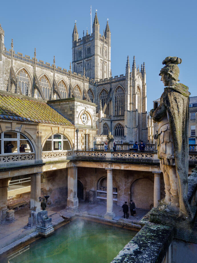 A large sculpture stands on top of roman baths ruins with an abbey beyond