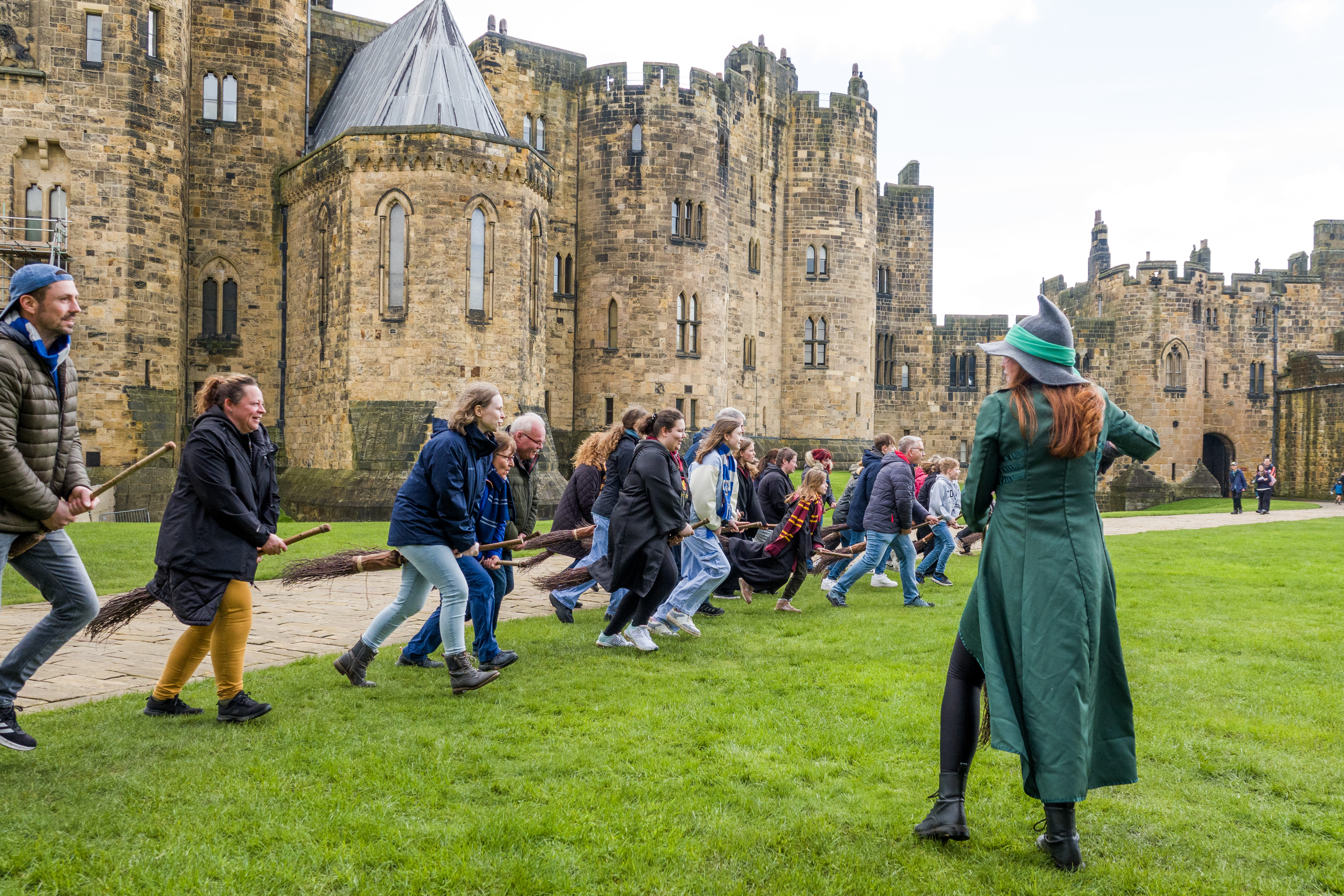 A group of people lined up for a Broomstick Training lesson with the wizarding professors by a large castle.