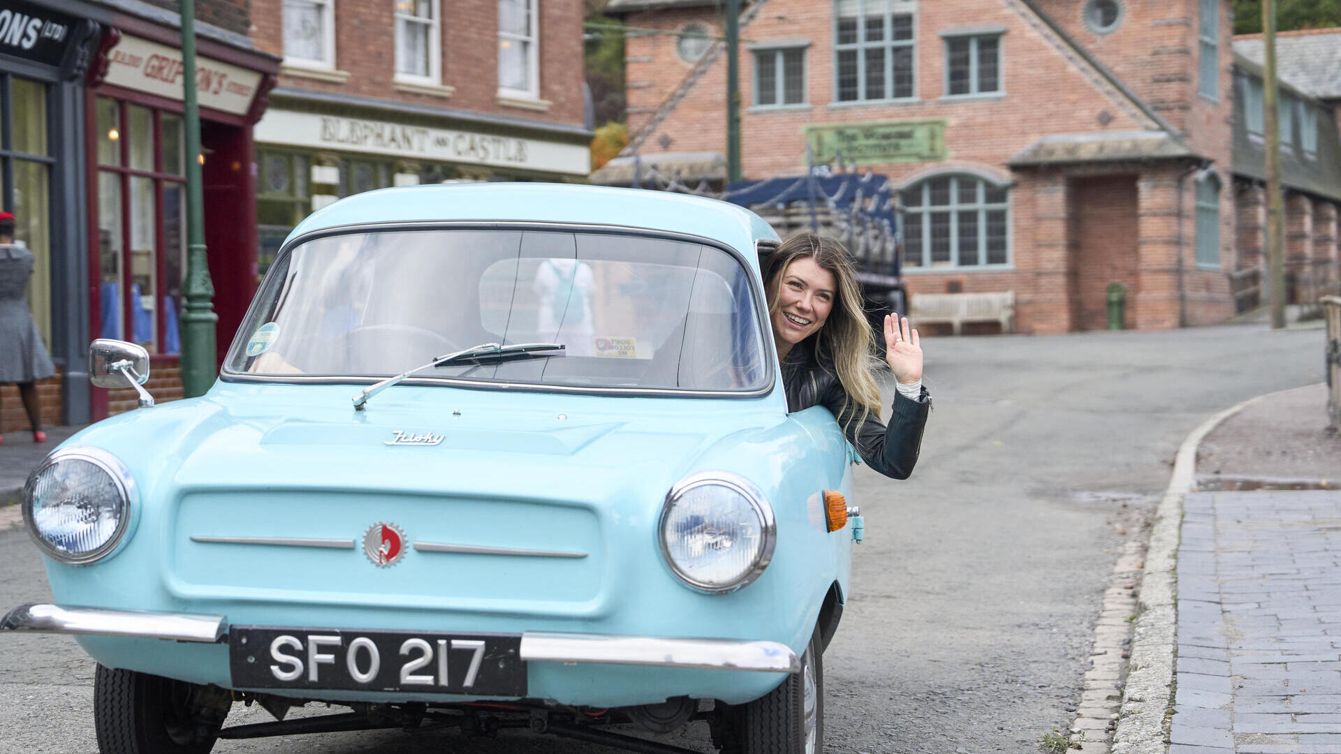 A woman waves from the window of a light blue vintage car on a quiet street with historic brick buildings and shops in the background.