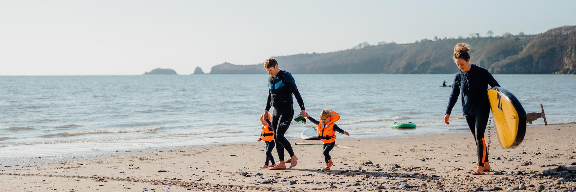 عائلة تمشي على الشاطئ، الرجل بصحبة طفلين صغيرين والمرأة تحمل لوح تزلج على شاطئ سوندرسفوت