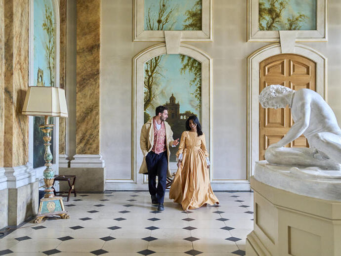 Elegant hall with classical decor, statue, ornate lamp, and two people in period attire walking and talking. Arched murals and wooden doors in the background.