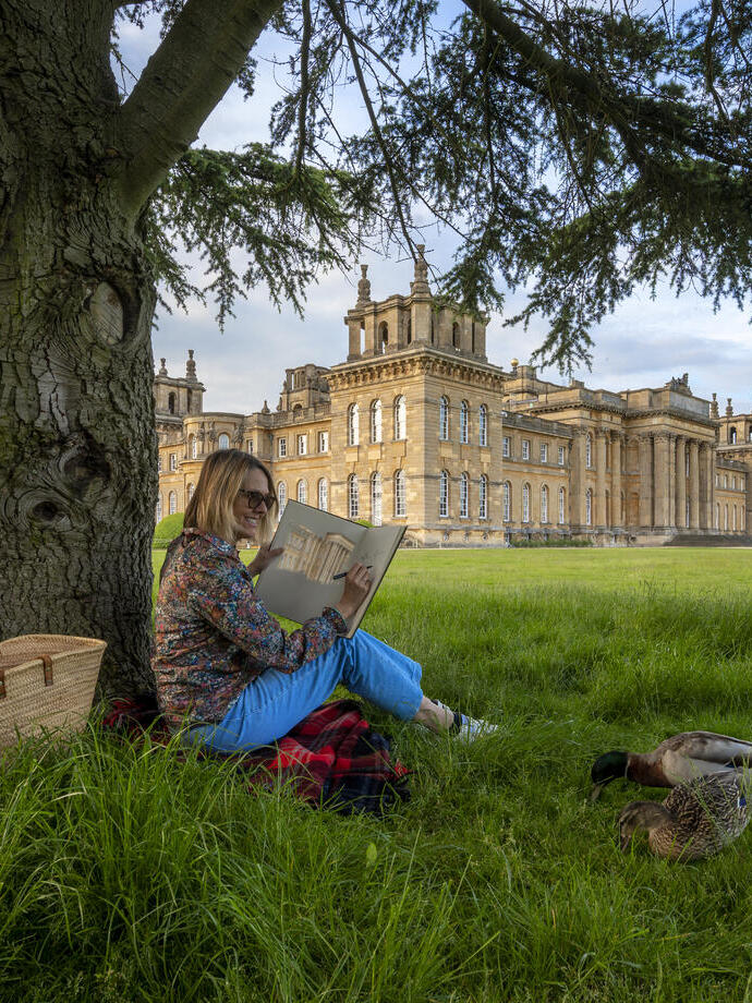 Woman sat under a tree sketching in the grounds of a large country estate