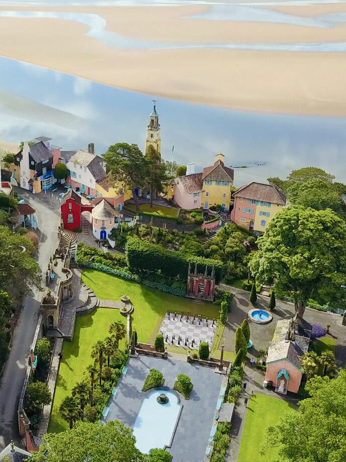 Aerial view of a colorful village with unique architecture, gardens, and a clock tower, set beside a sandy estuary and lush greenery.