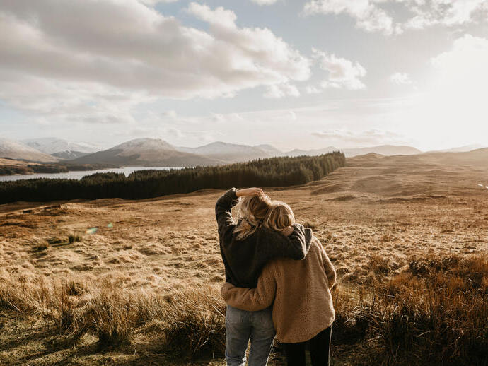 Zwei Freunde umarmen sich und blicken über das Tal auf einen See und die Berge