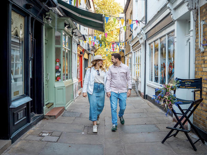 A couple walking and holding hands down a charming narrow street with colorful bunting overhead and shopfronts on either side.
