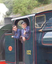 Two men wearing overalls standing in the cab of a vintage steam train.