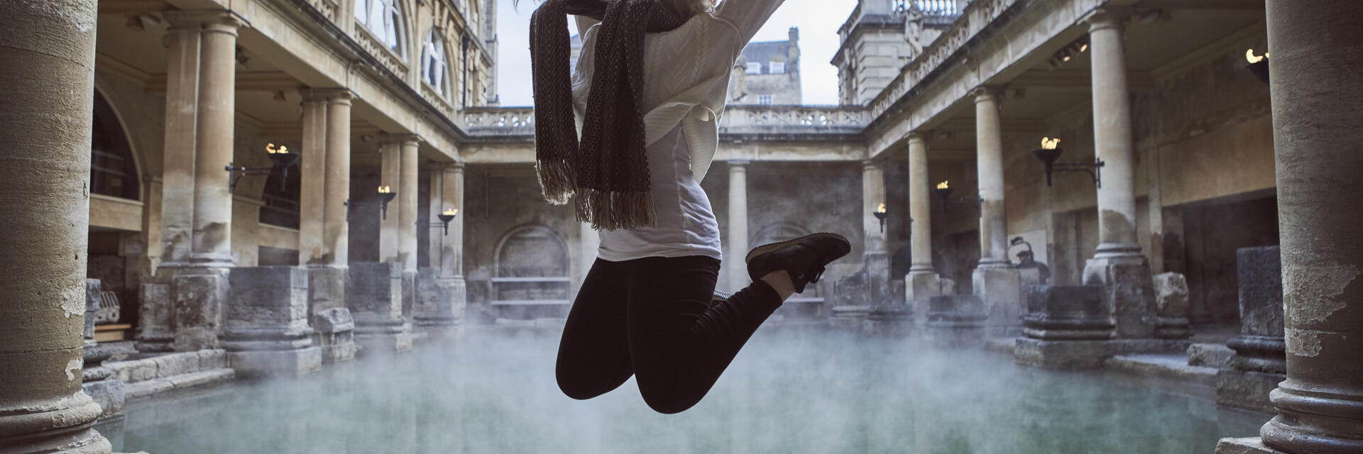 Frau springt in die Luft in einem römischen Freiluftbad, umgeben von historischen Gebäuden