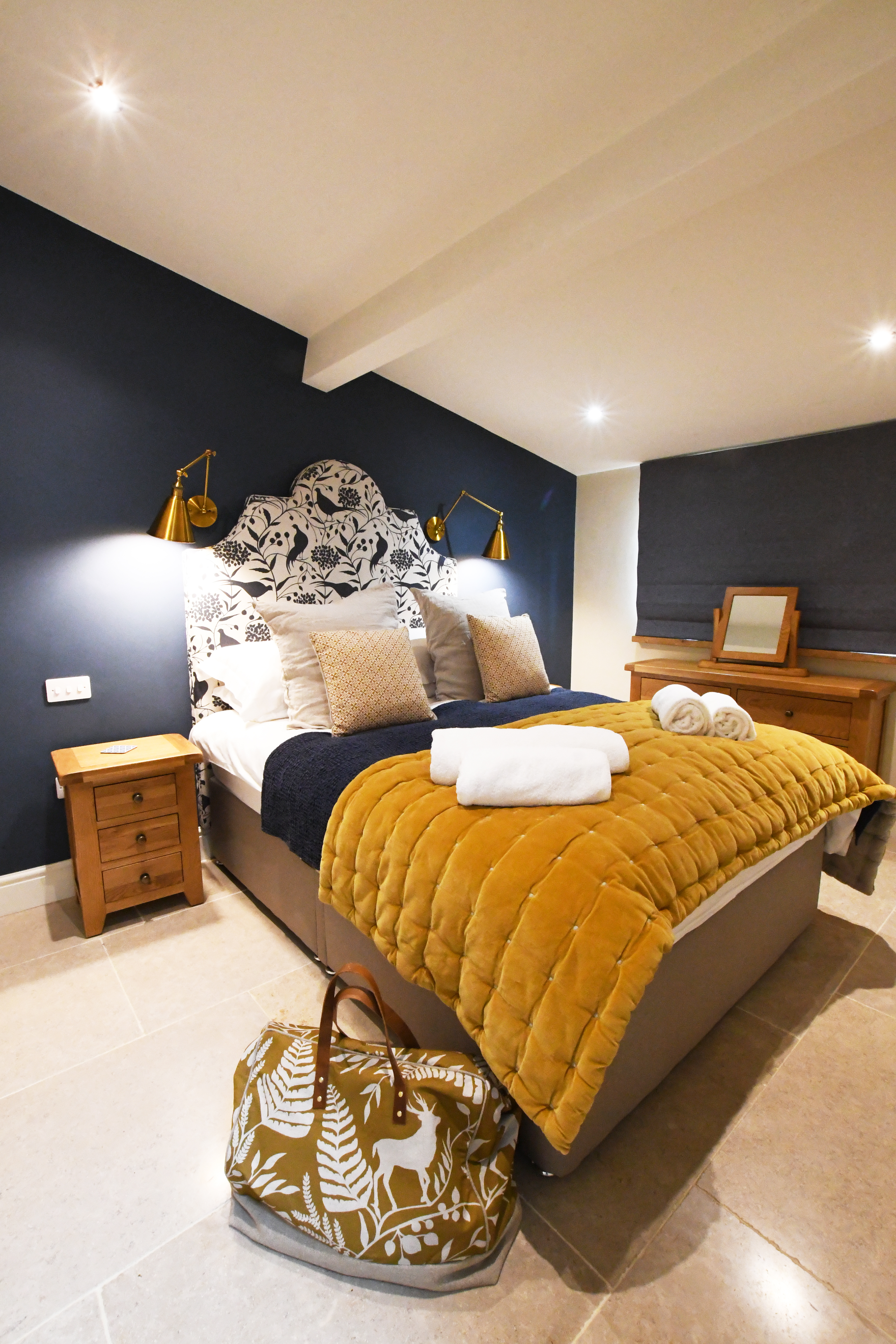 A double bedroom with dark blue and the bed has a mustard yellow padded beadspread. An over-night bag is positioned at the corner of the bed in the foreground.