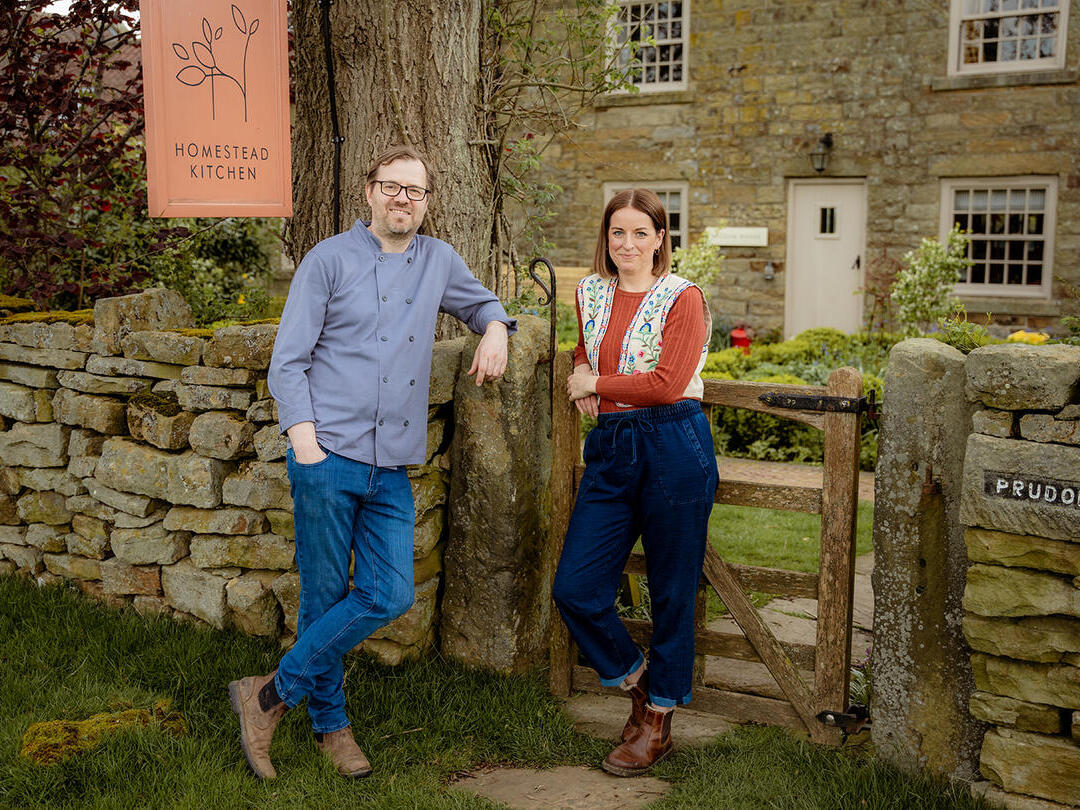 Two people standing in front of a countryside cottage. A sign reads: Homestead Kitchen