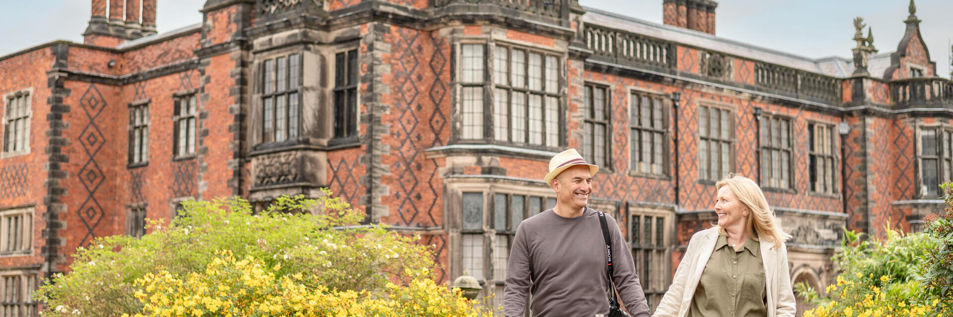 Hombre y mujer caminan juntos frente a una mansión histórica de ladrillo rojo y sonríen.