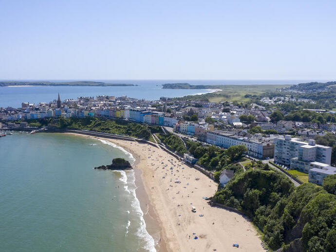 An overhead shot of a sandy beach lined with sunbathers, surrounded by brightly coloured townhouses and countryside.