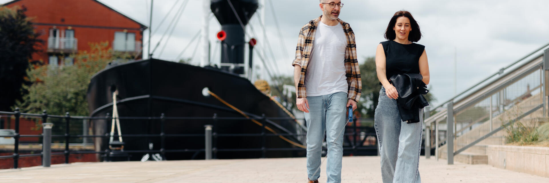 Two people walking a small dog along a paved waterfront with a historic ship and residential buildings in the background.