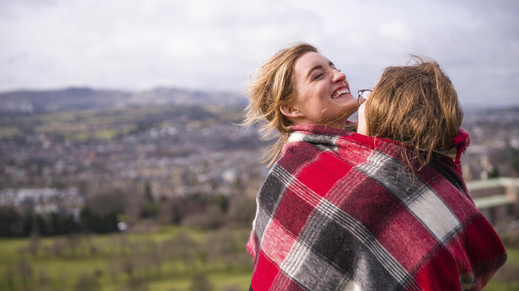 Two women wrapped in red checkered blanket standing on a hill overlooking a city