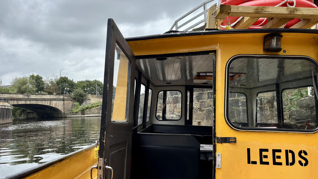 Yellow boat marked 'Leeds' on a river under a cloudy sky, with a stone bridge, greenery, and city buildings visible in the background.