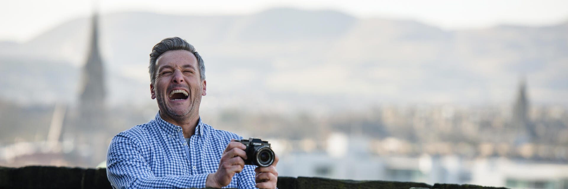 Smiling person holding a camera stands behind stone wall, with blurred cityscape and hills in the background.