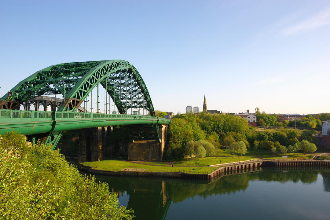 View of a green iron bridge crossing over a river, surrounded by trees and buildings