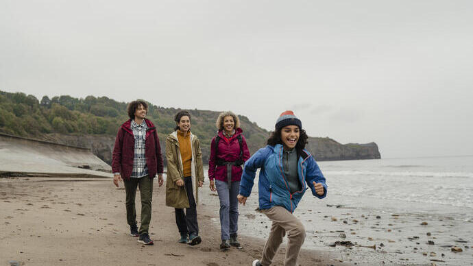 Group walking on a sandy beach with one child running ahead, cliffs and trees in the background under an overcast sky.