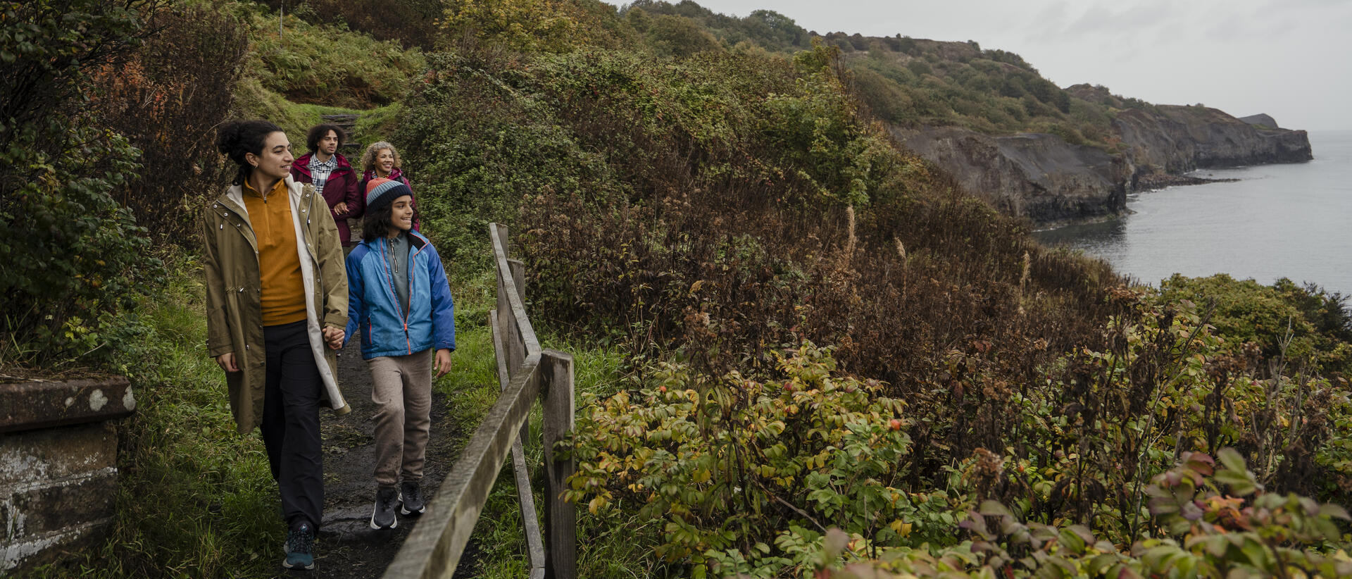 Group walking along a coastal path with lush greenery, cliffs, and sea in the background on a cloudy day.