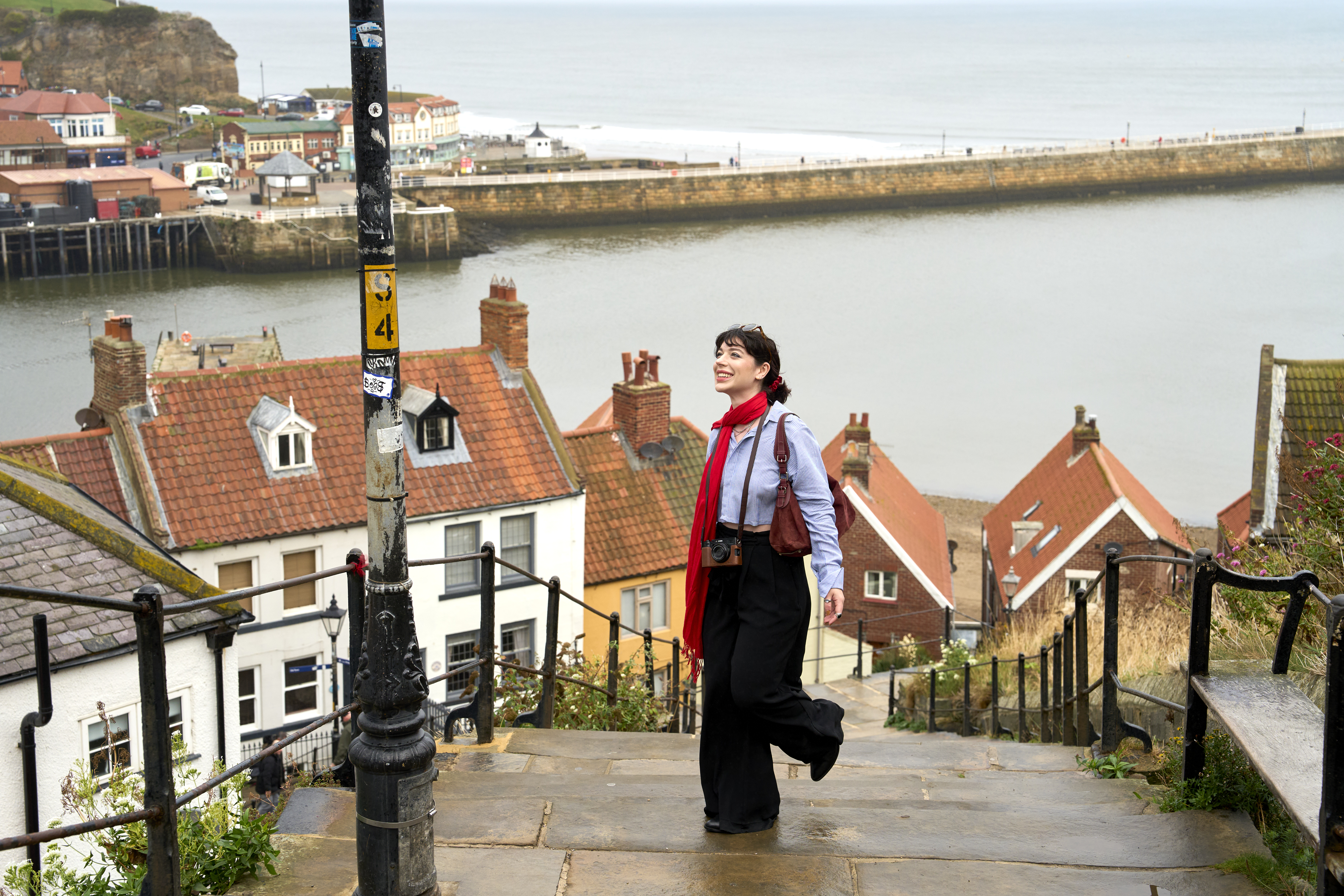Person walking up stone steps by red-roofed houses overlooking a river and seaside harbor, with cliffs and coastline in the background.