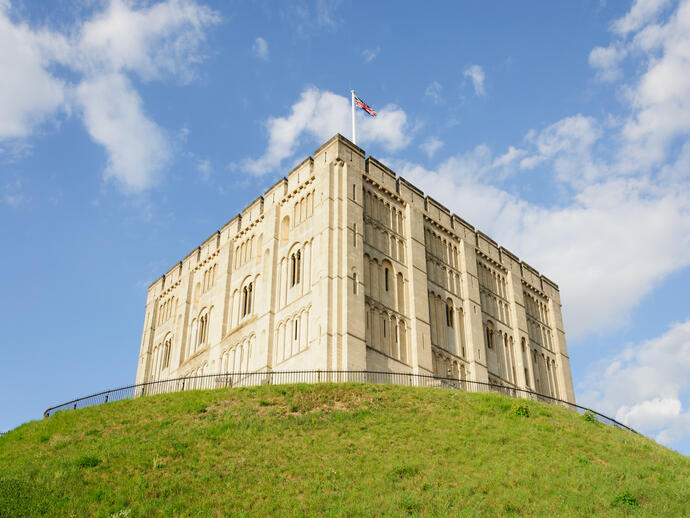A large stone castle sits atop a grassy hill under a blue sky with scattered clouds, a flag flying above the building.