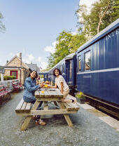 Two people dining at a picnic table on a historic train station platform with a blue train and Bassenthwaite Lake station sign visible.