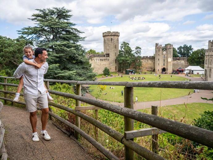 Hombre lleva a una niña sobre sus hombros caminando por un sendero junto a un castillo histórico, con personas sentadas en el césped al fondo.
