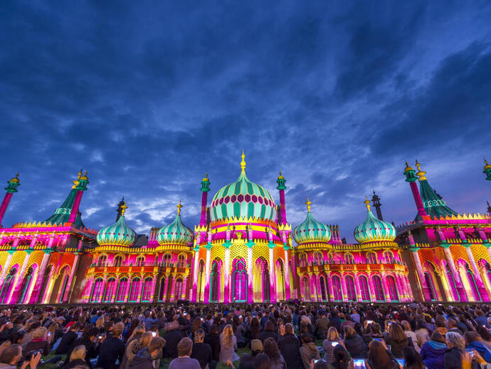 Crowd sitting on the ground in front of a large ornate building lit up with bright colours