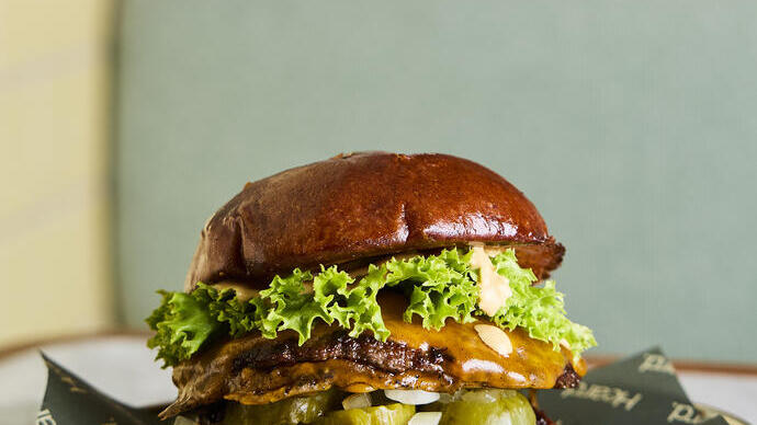 Close-up of a gourmet burger with lettuce, cheese, pickles, onions, and sauce in a glossy brioche bun on a plate in a restaurant setting.