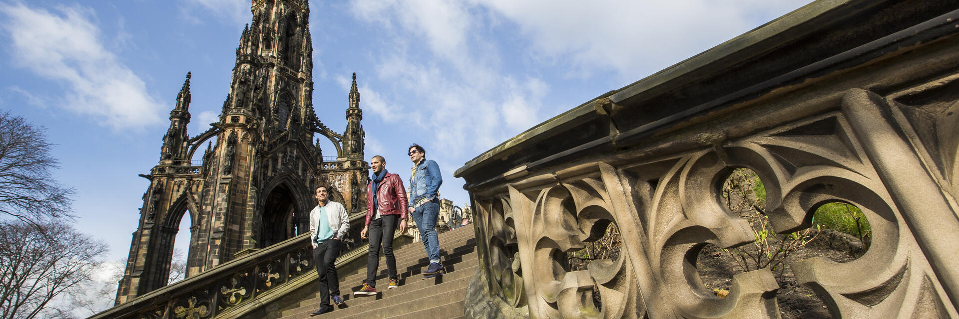 Three people walking down stone steps near the ornate Scott Monument under a partly cloudy blue sky in Edinburgh.
