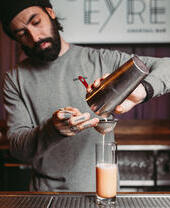 Un barista prepara un cocktail in una rilassante sala ristorazione all'aperto con una vasta gamma di venditori che offrono snack locali e internazionali, oltre a bevande artigianali.