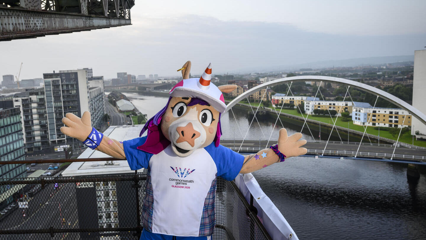 Mascot in costume stands on a bridge overlooking a river, modern buildings, and an arched bridge in a cityscape.