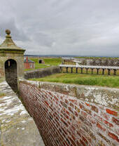 A brick defensive wall with a small tower amid a larger stone fortification under grey skies.