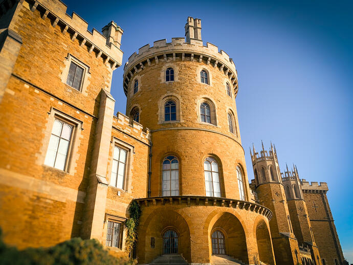 A large historic castle with rounded and square towers, arched windows, and golden stone walls under a clear blue sky.