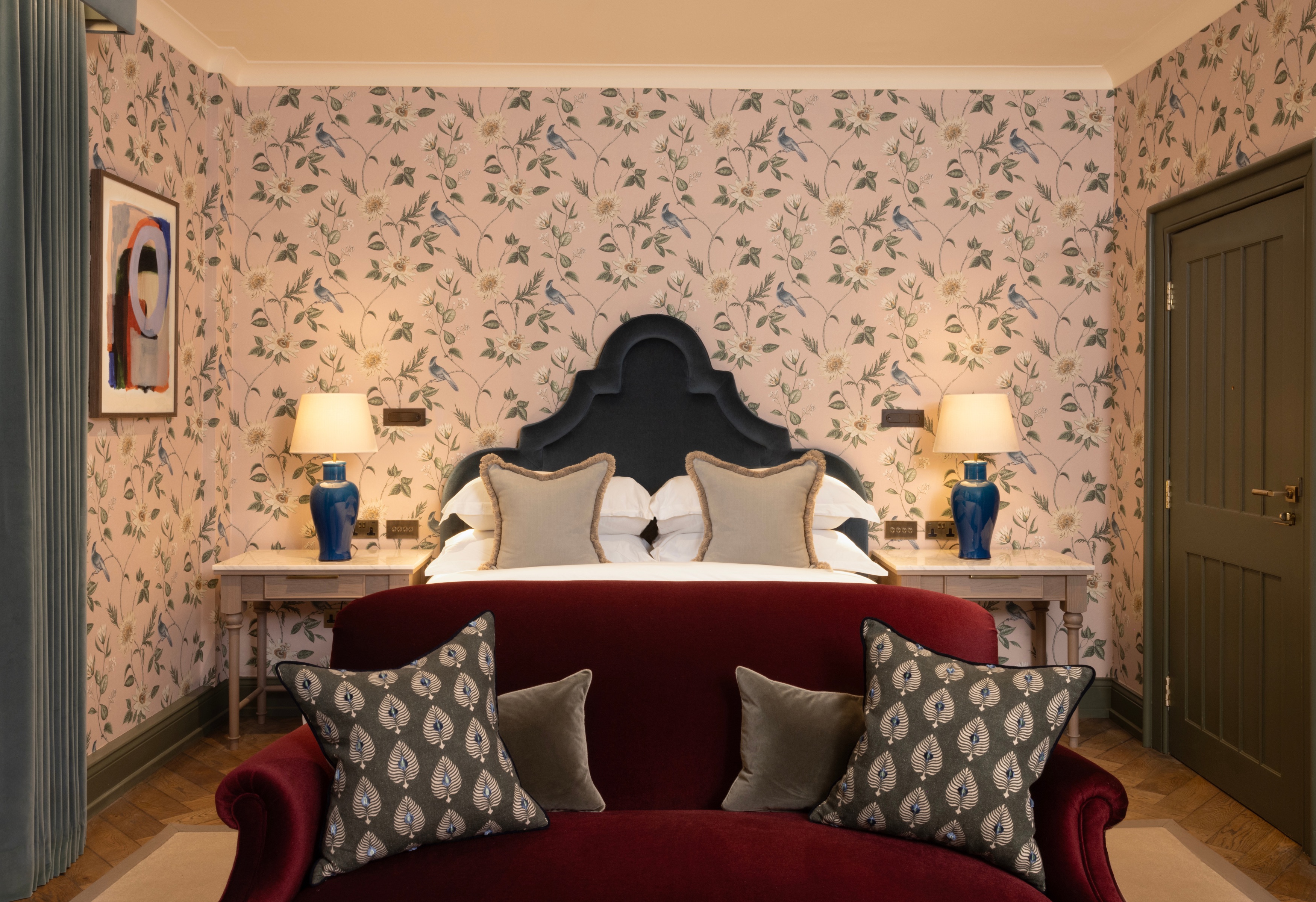 Luxuriöses Hotelzimmer mit rotem Samtsofa am Fußende des Bettes und blumiger Tapete an den Wänden dahinter.