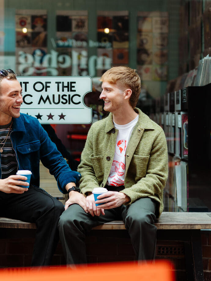 Zwei Männer sitzen mit Getränken zum Mitnehmen vor einem Laden