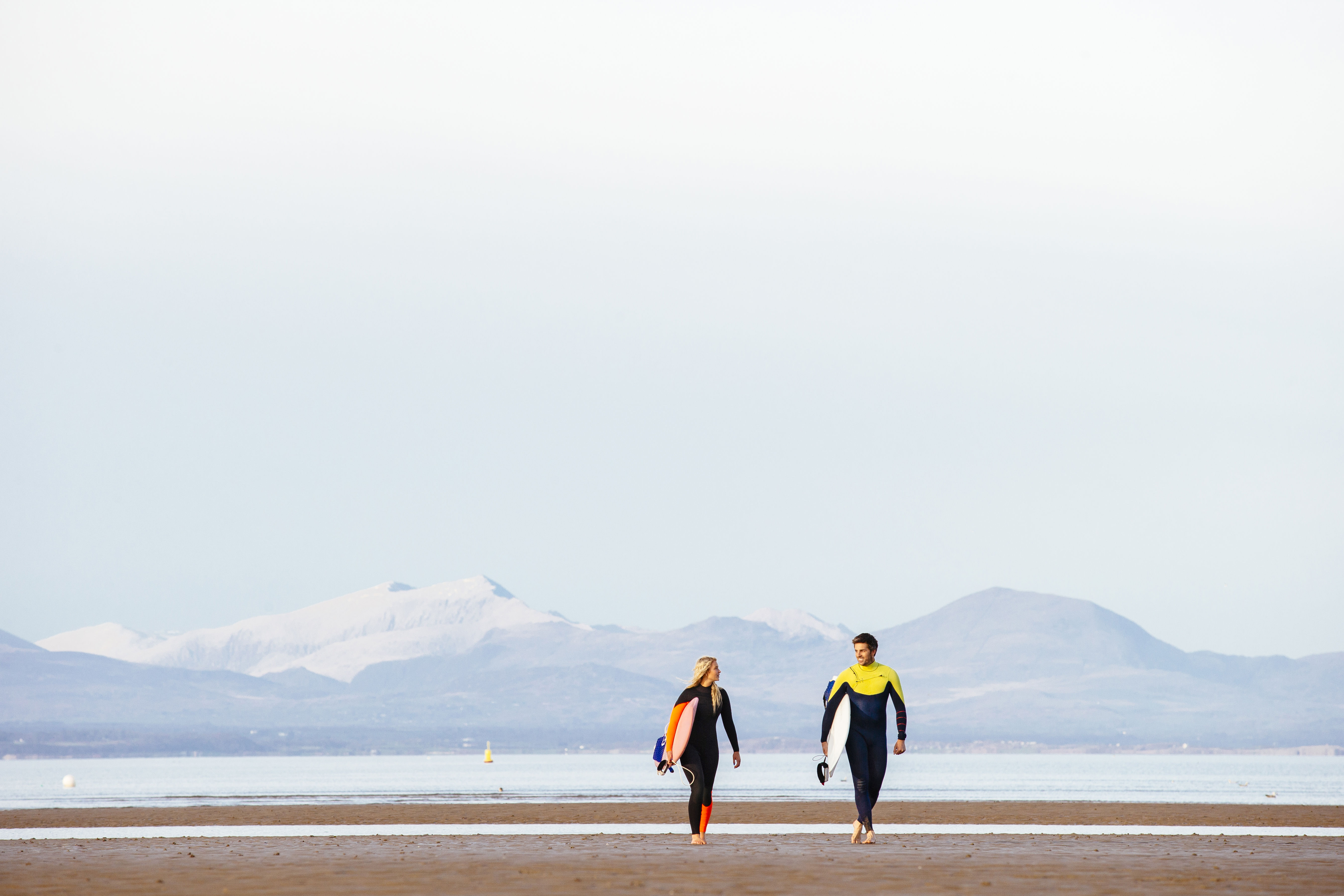 Man and woman, in wetsuits, carrying surfboards on beach