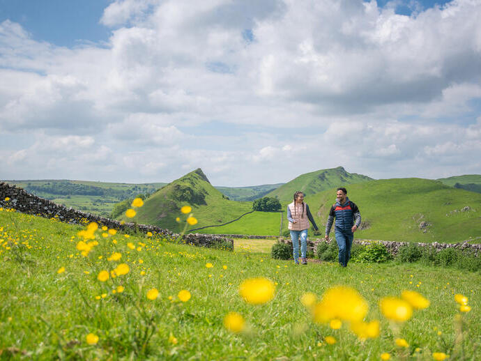 Two people walking through a green meadow with yellow flowers, hills, and stone walls under a partly cloudy sky.