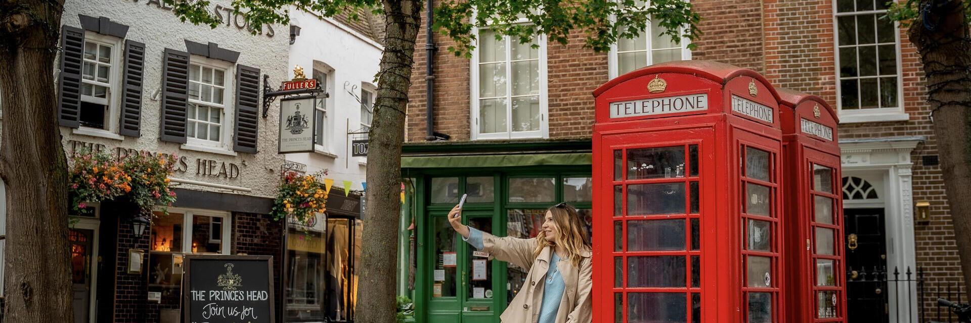 Una mujer apoyada en una cabina telefónica roja y haciéndose un selfie
