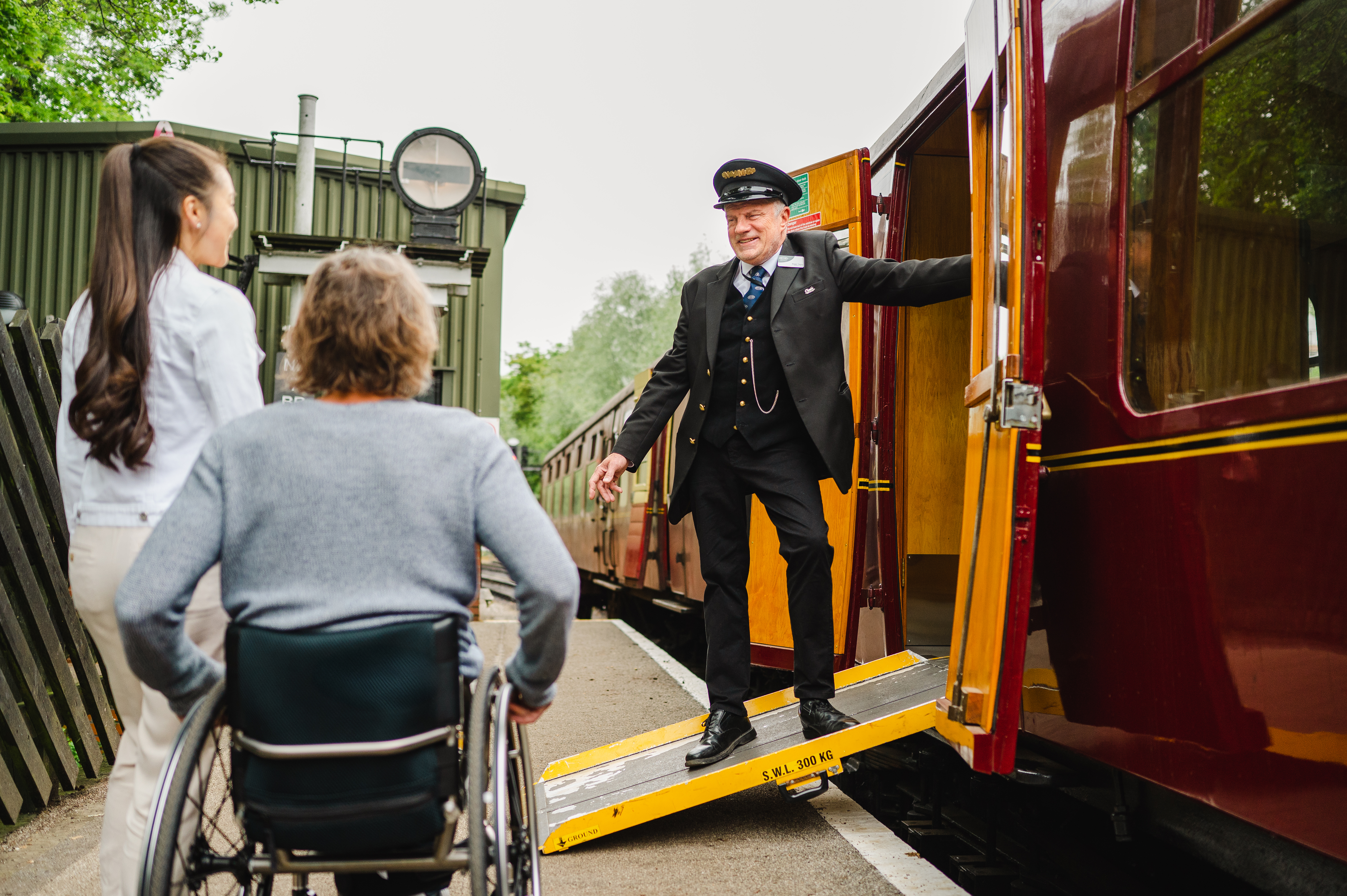 Guard stands on a ramps and leans out of heritage train carriage greeting woman and man using a wheelchair