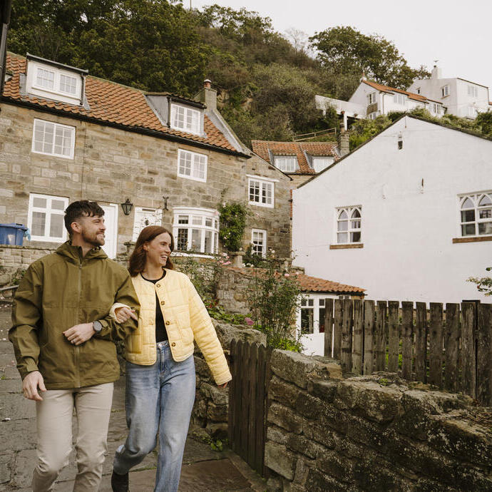 Couple walking on a cobblestone street in a quaint village with stone houses, red-tiled roofs, greenery, and hillside cottages in the background.